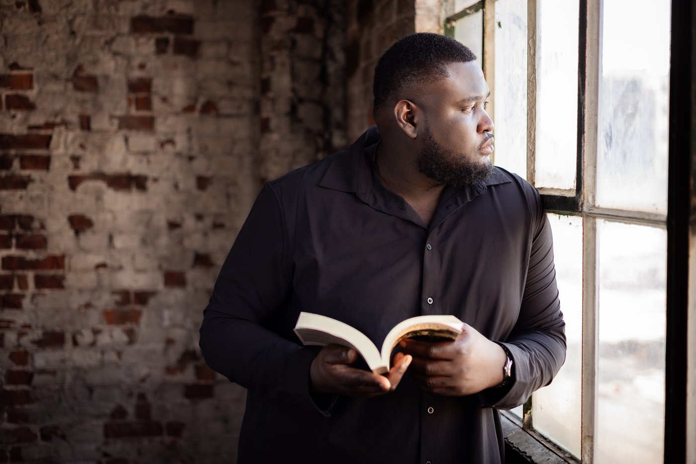 Christian Hakiza standing by a large window, holding an open book and looking outside