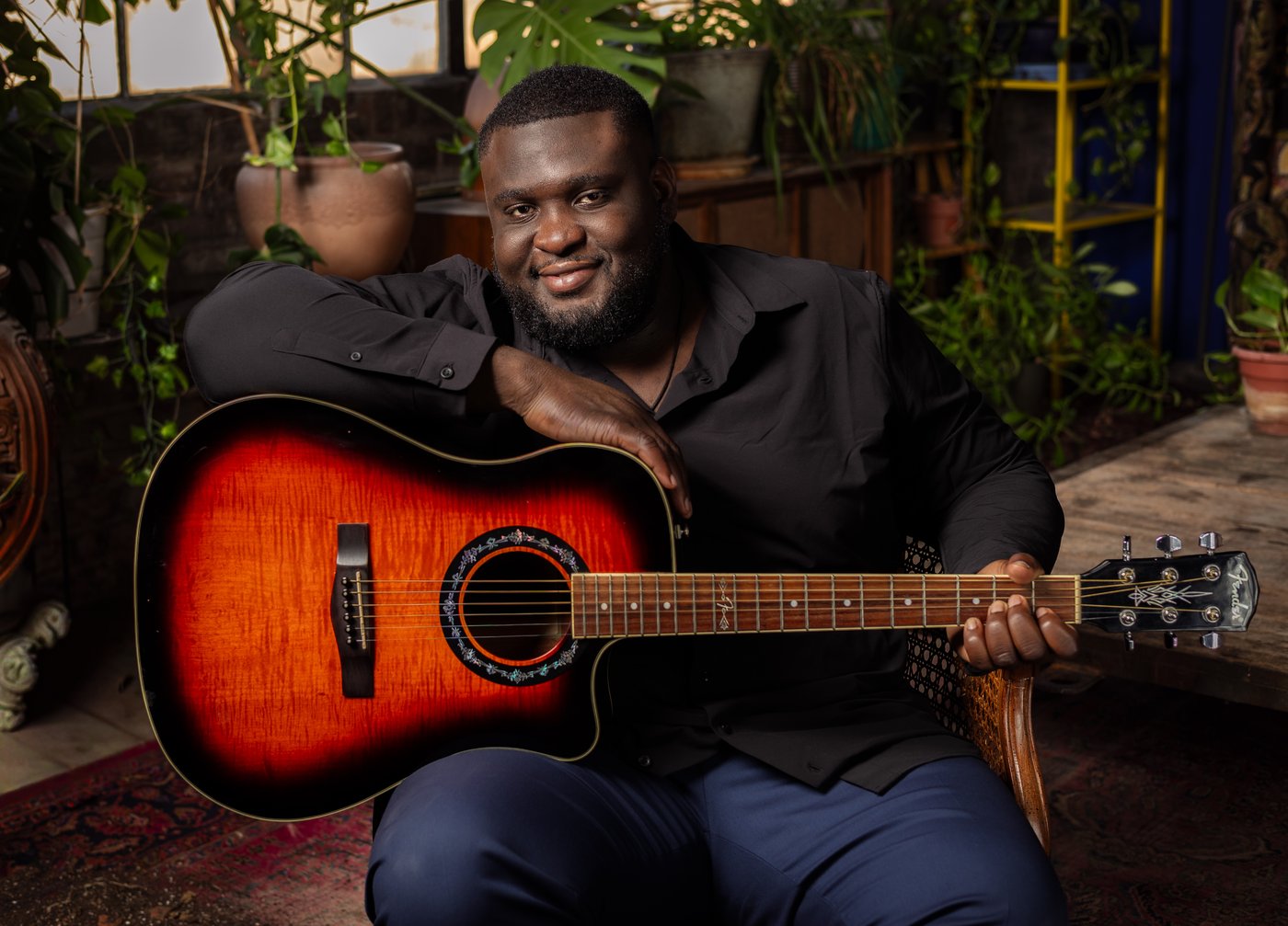 Christian Hakiza smiling at the camera, holding an acoustic guitar in a room full of plants